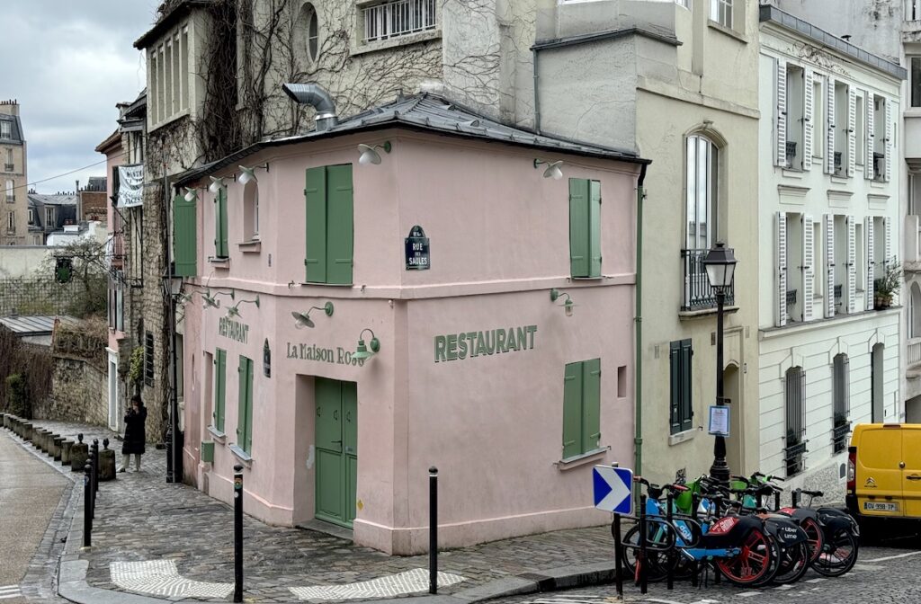 Pink cafe with greens shutters on corner of cobblestoned street with historic buildings and bikes in background. Montmartre, Paris, France.