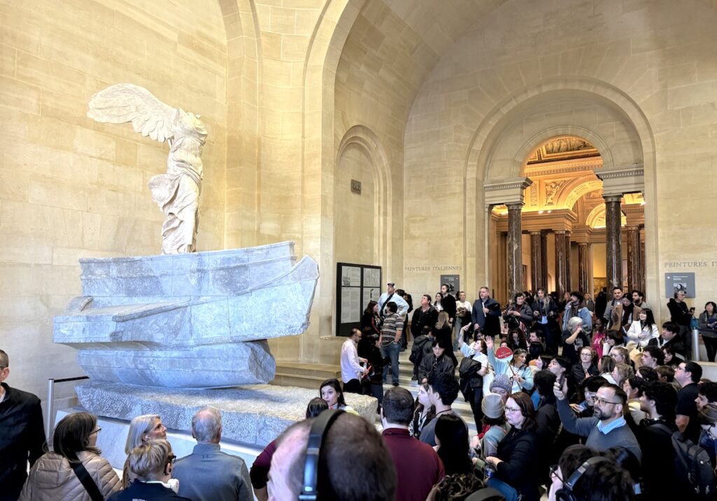 Large marble statue with wings on boat surrounded by huge crowd in spacious museum. Winged Vicotry of Samothrace (Nike of Samothrace), Louvre Museum, Paris, France.