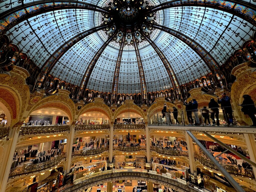 Circular shopping mall with huge, domed stain glass ceiling and open architecture with elaborate gold details. Galeries Lafayette department store, Paris, France.