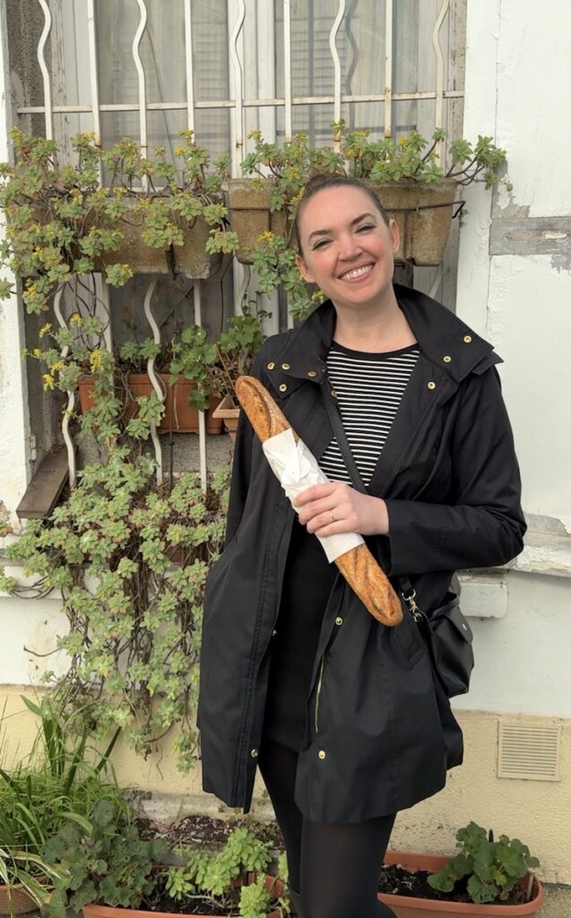 Woman smiling and holding French baguette in front of courtyard garden. Paris, France.