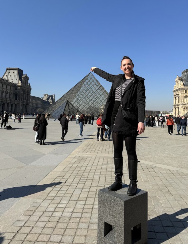 Woman smiling and pointing and triangle glass building at Louvre, Paris, France.