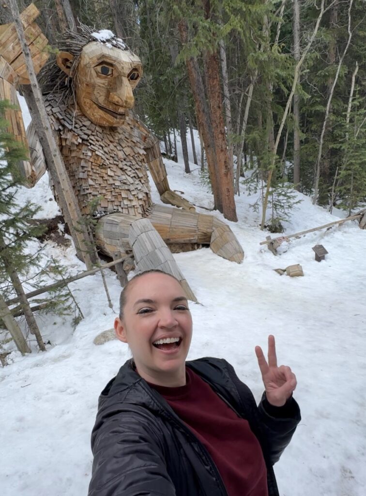 Woman smiling and taking a selfie in front of 15-foot tall troll made of reclaimed wood in snowy pine tree forest. Isak Heartstone troll, Breckenridge, Colorado in spring.