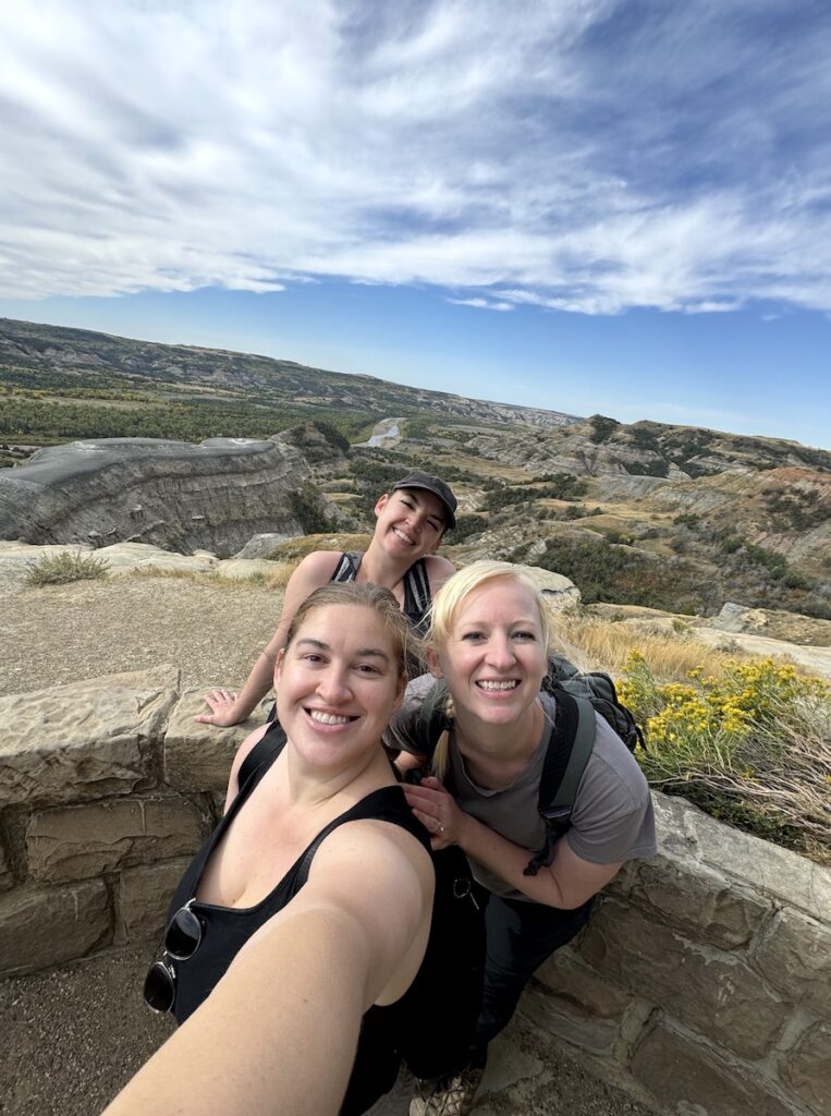 Women smiling at camera with mountain and badland views in background on sunny day. River Bend Overlook, Theodore Roosevelt National Park North Unit, North Dakota.