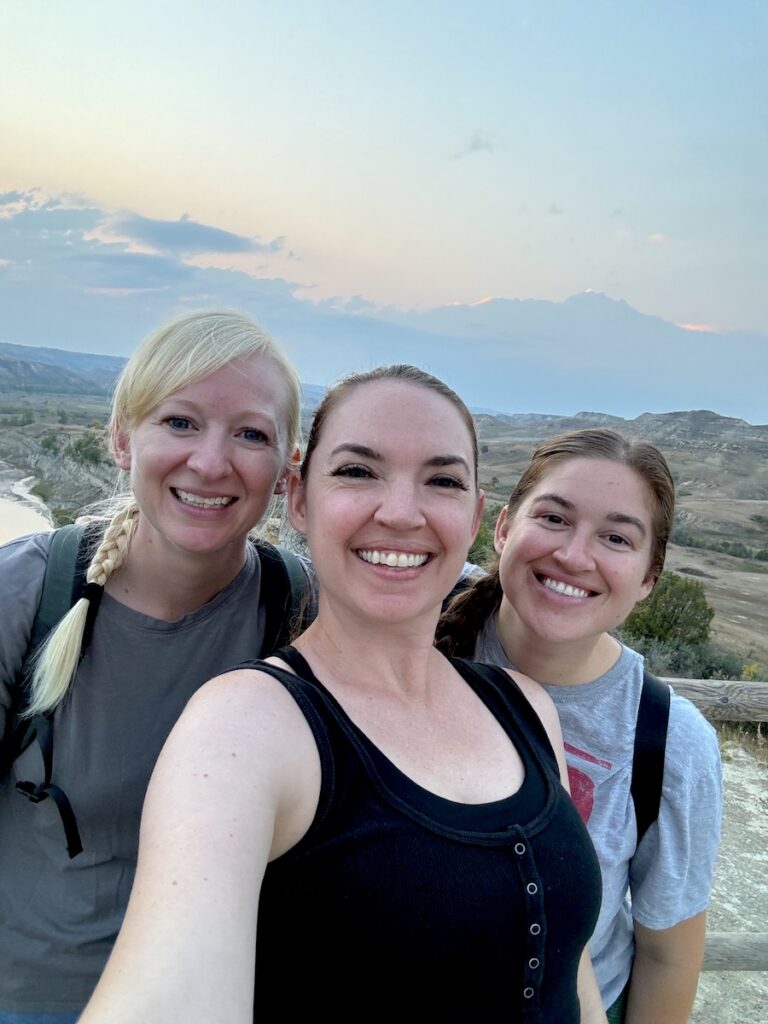 Three women smiling at camera with sunset in background on hiking trail, Wind Canyon Trail, Theodore Roosevelt National Park South Unit, North Dakota.