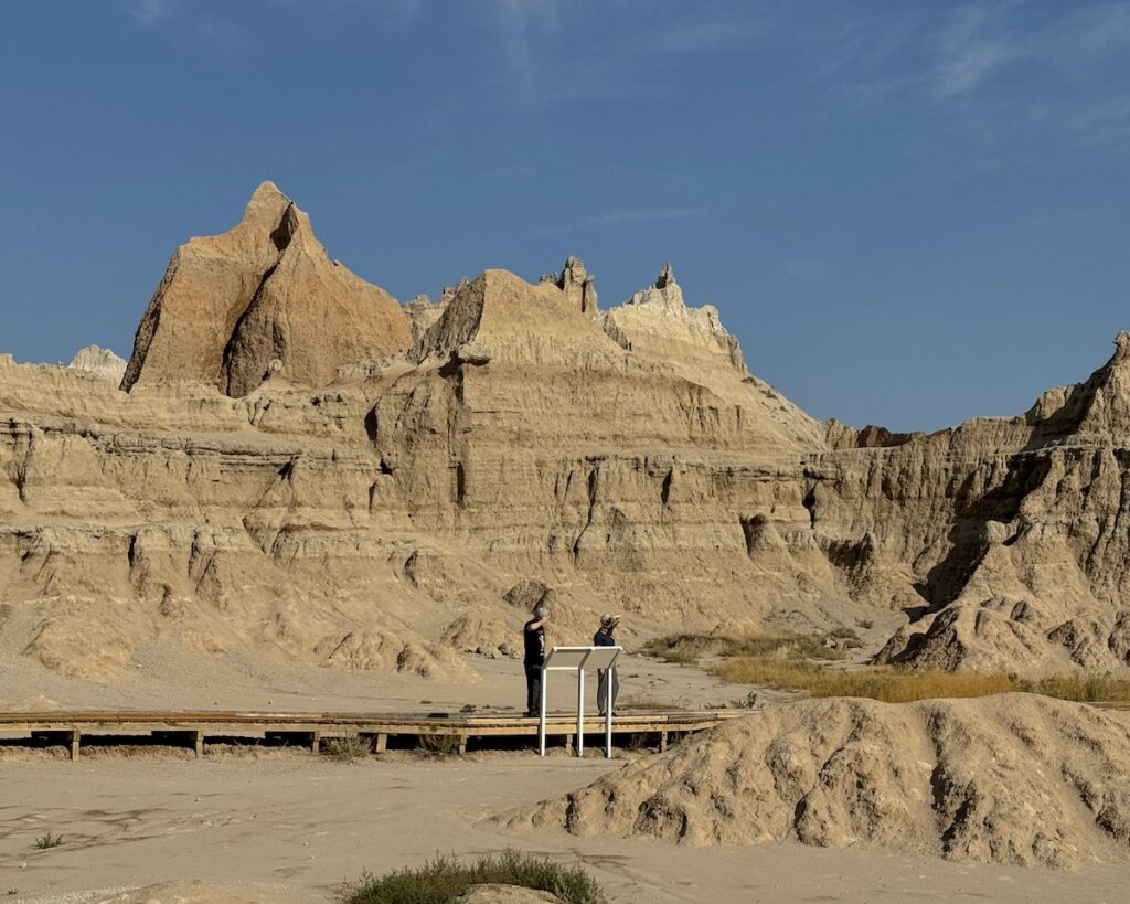 Boardwalk trail with people looking at infographics with badland canyons rising up in background on sunny day. Fossil Exhibit Trail, Badlands National Park, South Dakota.