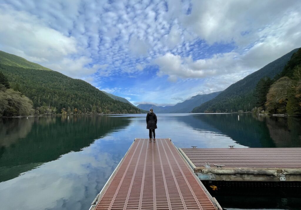 Woman standing on edge of dock overlooking very calm lake surrounded by mountain views on sunny fall afternoon. Crescent Lake, Olympic National Park, Olympic Peninsula, Washington.