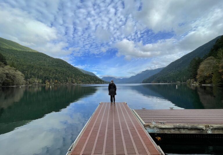 Woman standing on edge of dock overlooking very calm lake surrounded by mountain views on sunny fall afternoon. Crescent Lake, Olympic National Park, Olympic Peninsula, Washington.