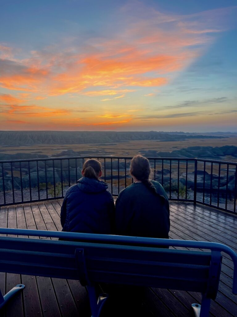 Two women sitting on bench watching sun rise over the colorful badlands. Sunrise at Big Badlands Overlook, Badlands National Park, South Dakota.