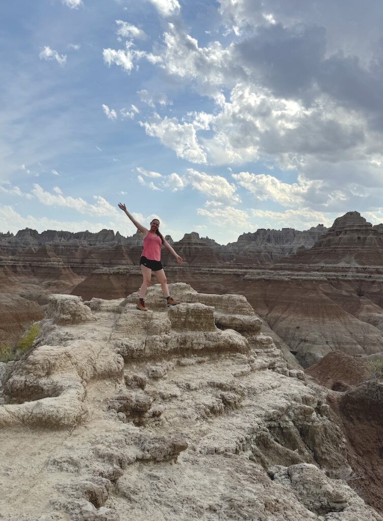 Woman posing on jagged rock surrounded by colorful badlands landscape on sunny afternoon. Door Trail, Badlands National Park, South Dakota.