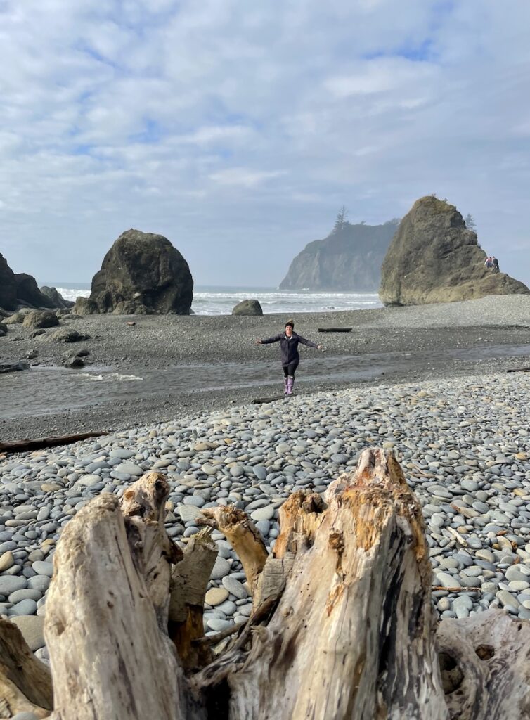 Woman walking on rocky beach with sea stacks rocky shore in background on sunny afternoon. Ruby Beach, Olympic National Park, Olympic Peninsula, Washington.