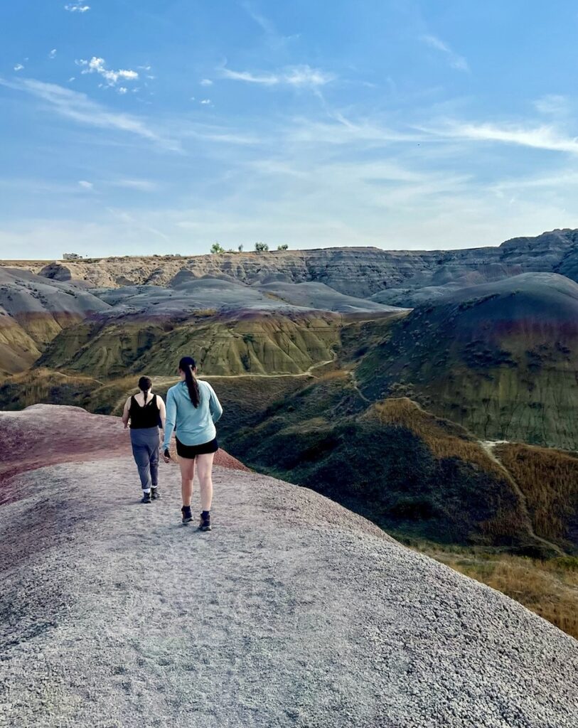 Two women walking on colorful badlands with yellow and purple badlands in background on sunny afternoon. Yellow Mounds Overlook, Badlands National Park, South Dakota.