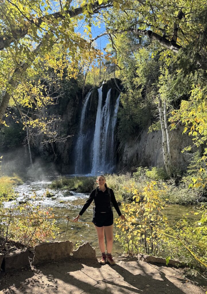 Woman smiling in front of tall single-drop waterfall framed by yellow trees on sunny day in early fall. Spearfish Falls, Spearfish Canyon Byway, Black Hills, South Dakota.