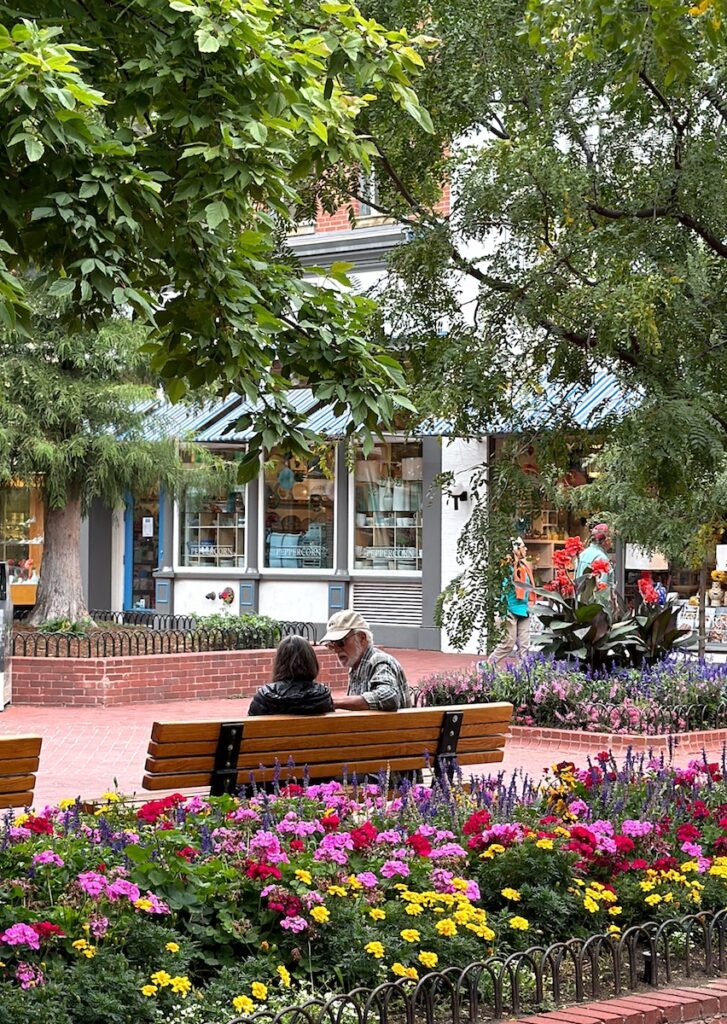 Couple sitting on bench of Pearl Street Mall with brick sidewalk, flowers, and green trees. Pearl Street Mall, Boulder, Colorado.