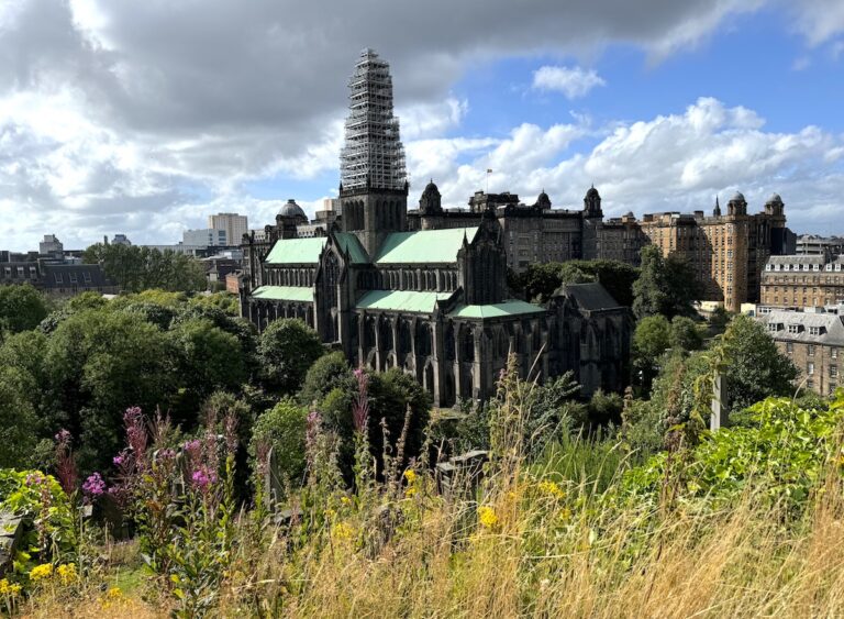 Dramatic stone cathedral with green roof and old city buildings in background with flowers and greenery in foreground on sunny day. Glasgow Cathedral, Glasgow, Scotland, United Kingdom.