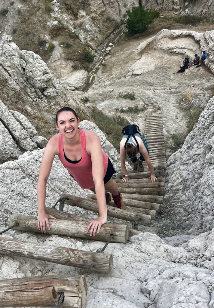 Two women climbing wooden rung ladder up badland rock formations. Notch Trail, Badlands National Park, South Dakota.