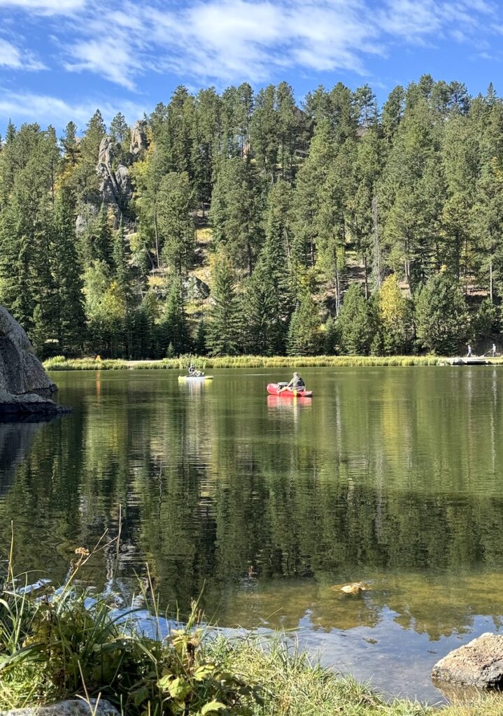 People kayaking on calm lake with mountains and pine trees in background on sunny day. Sylvan Lake, Custer State Park, Black Hills, South Dakota.