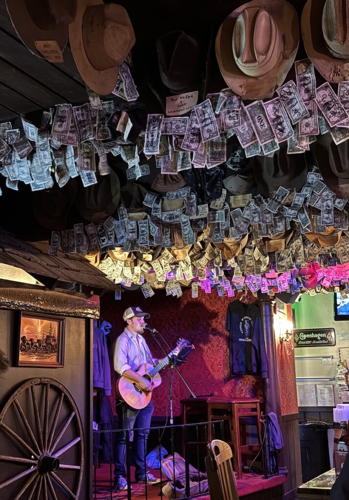 Man singing and playing guitar in small country bar with dollar bills and country hats on ceiling. Little Missouri Saloon, Medora, North Dakota.