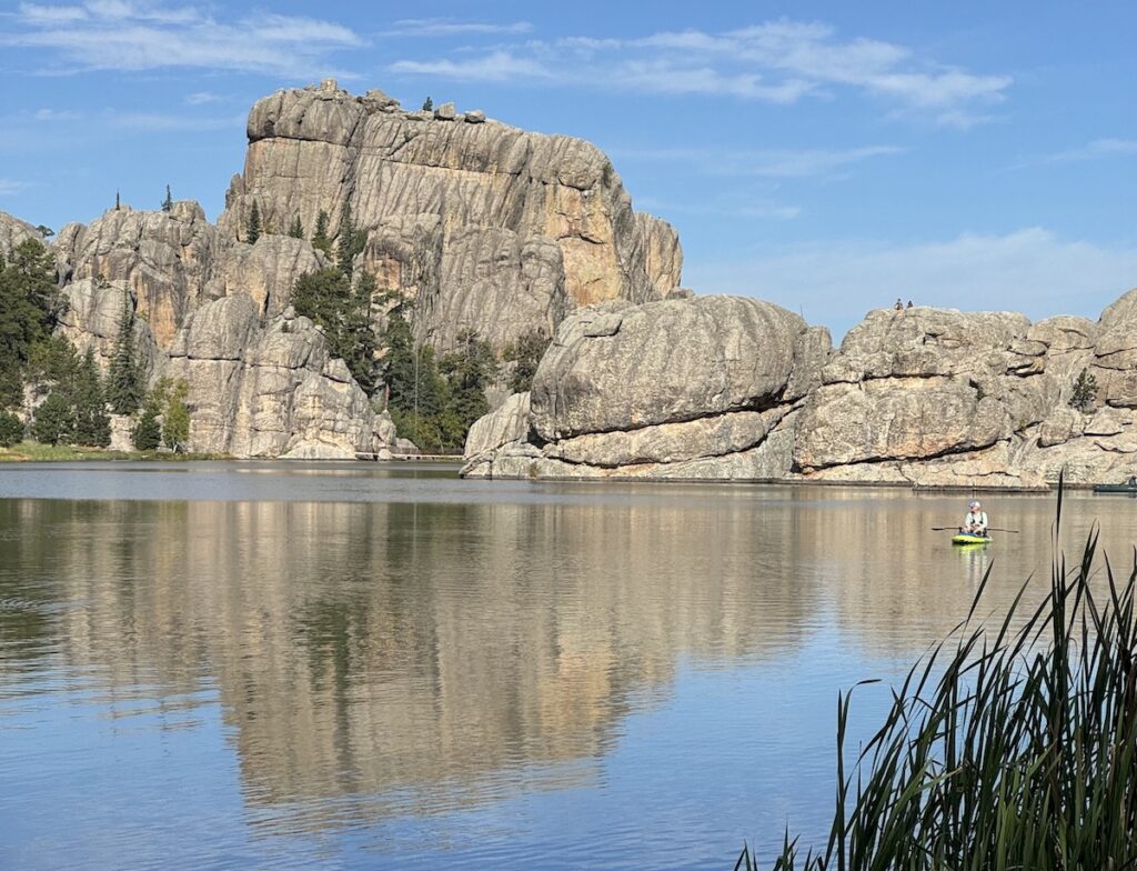 Person kayaking on calm lake with huge, dramatic granite boulders in background on sunny day. Sylvan Lake, Custer State Park, Black Hills, South Dakota.