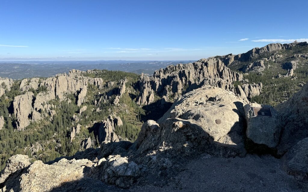 Jagged granite mountains and boulders peaking out from lush green forest of pine trees with blue skies in background. View from summit of Little Devils Tower hiking trail, Custer State Park, Black Hills, South Dakota.