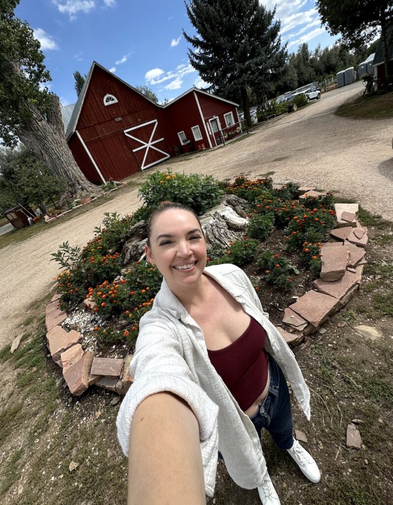 Woman smiling at camera with with red barn and flowers in background on sunny fall day. Ya Ya Farm & Orchard, Longmont, Colorado.