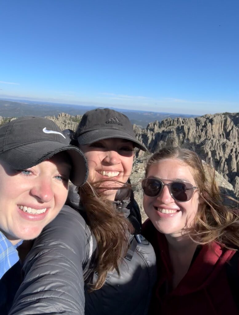 Three woman smiling at camera from summit of Little Devils Tower hiking trail with granite mountains and blue skies in background. Little Devils Tower, Custer State Park, Black Hills, South Dakota.