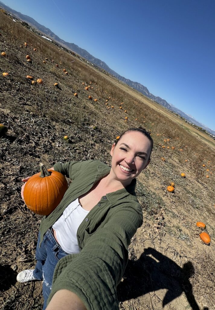 Woman smiling and holding pumpkin at u-pick pumpkin patch with flatiron and mountain views in background on sunny fall day. Rock Creek Farms, Broomfield, Colorado.