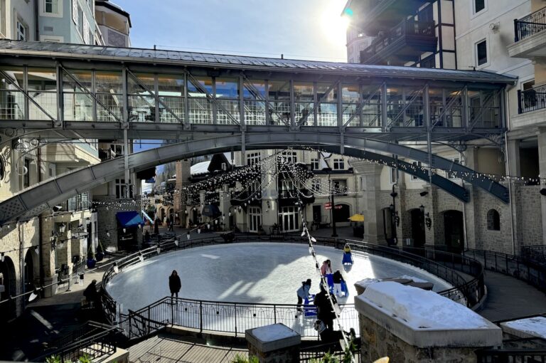 Charming ice skating rink under string lights and pedestrian bridge, surrounded by luxury hotel with Bavarian-architecture on sunny afternoon during Christmas. The Rink at Arrabelle, Lionshead Village, Vail, Colorado.