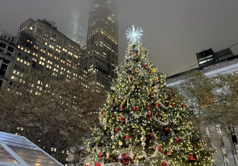 Colorful Christmas tree lit up at night with skyscraper buildings rising up behind it on cloudy gloomy night. Bryant Park Winter Village, Midtown Manhattan, New York City, New York.
