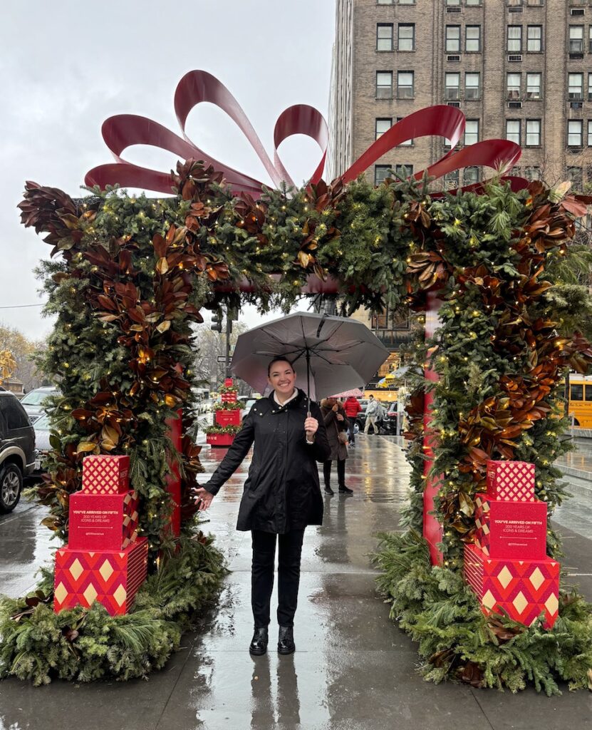Woman smiling and holding umbrella under large garland Christmas decor on rainy afternoon on 5th Ave. Fifth Avenue Christmas displays, Midtown Manhattan, New York City, New York.