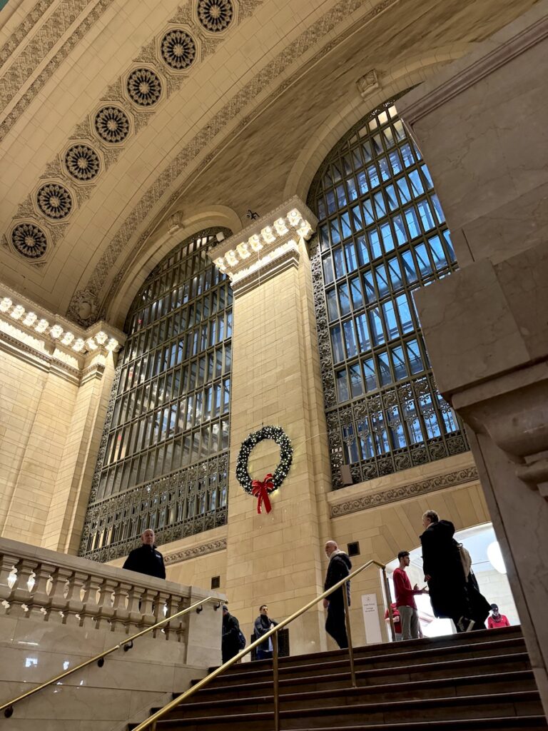 Interior of train station with oversized windows, elaborate ceiling art, and lit Christmas wreath. Grand Central Station Terminal, Midtown Manhattan, New York City, New York.