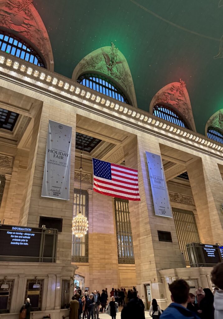 Interior of train station with oversized windows, elaborate ceiling art lit up in green and red, and a large American flag. Grand Central Station Terminal, Midtown Manhattan, New York City, New York.