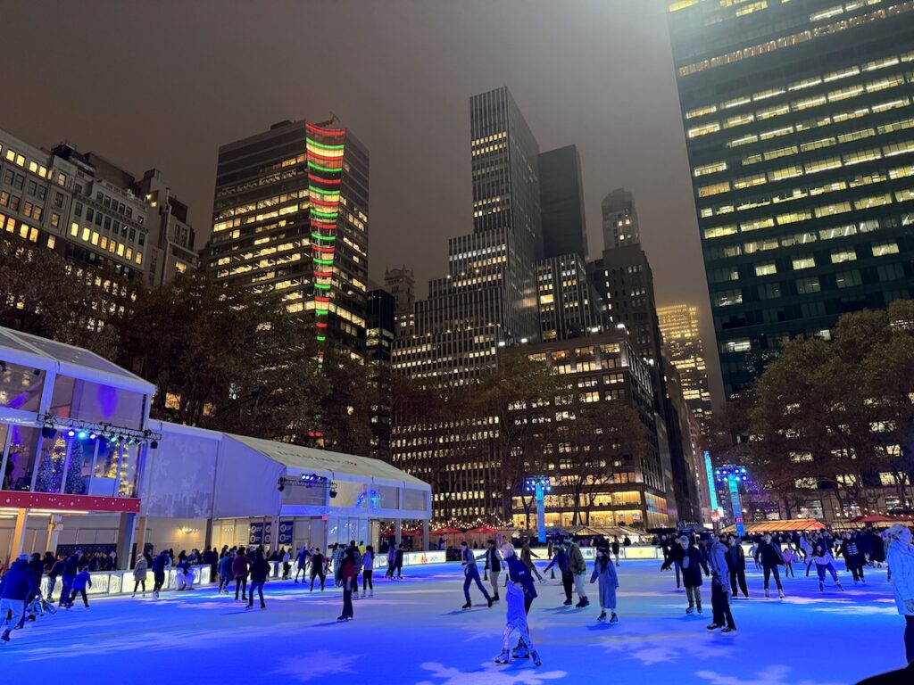 Ice skating rink full of people surrounded by marketplace pop-ups and huge towering buildings at night. Bryant Park Winter Village, Midtown Manhattan, New York City, New York.
