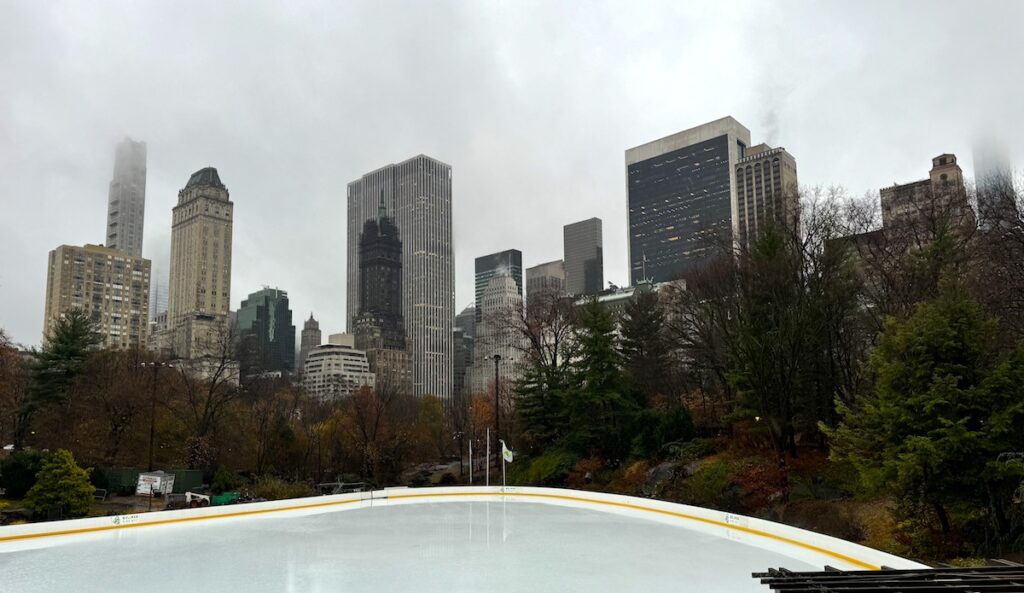 Empty ice skating rink surrounding by trees with large skyscraper buildings in background on gloomy and rainy afternoon. Wollman Rink, Central Park, Manhattan, New York City, New York.