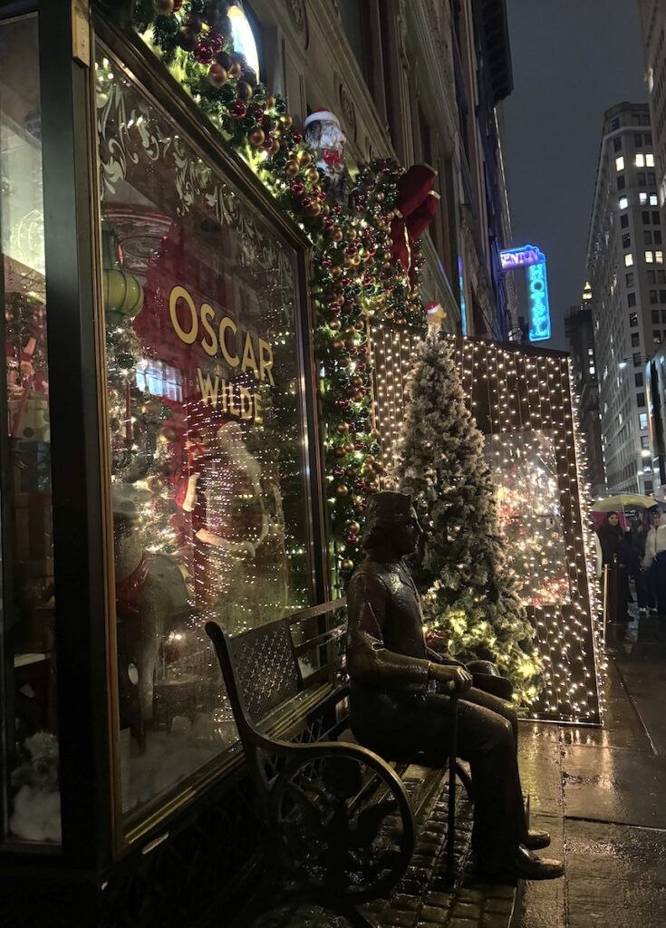 Exterior of bar covered in Christmas decorations at night with bronze statue of a man sitting on a bench. Oscar Wilde - Christmas bar, Manhattan, New York City, New York.
