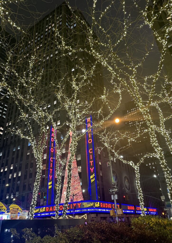 Christmas lights and Christmas decor on tall buildings lit up at night, neon signs read "Radio City." Radio City Music Hall, Midtown Manhattan, New York City, New York.