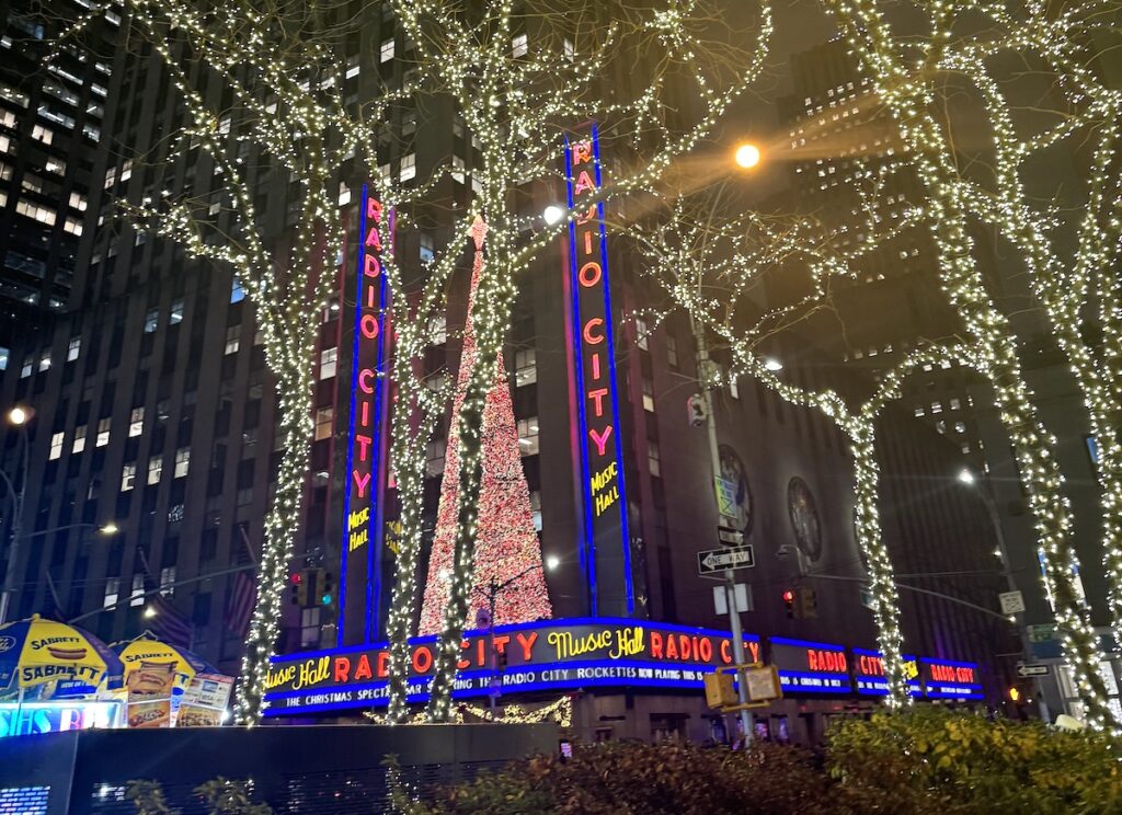 Christmas lights and Christmas decor on tall buildings lit up at night, neon signs read "Radio City." Radio City Music Hall, Midtown Manhattan, New York City, New York.