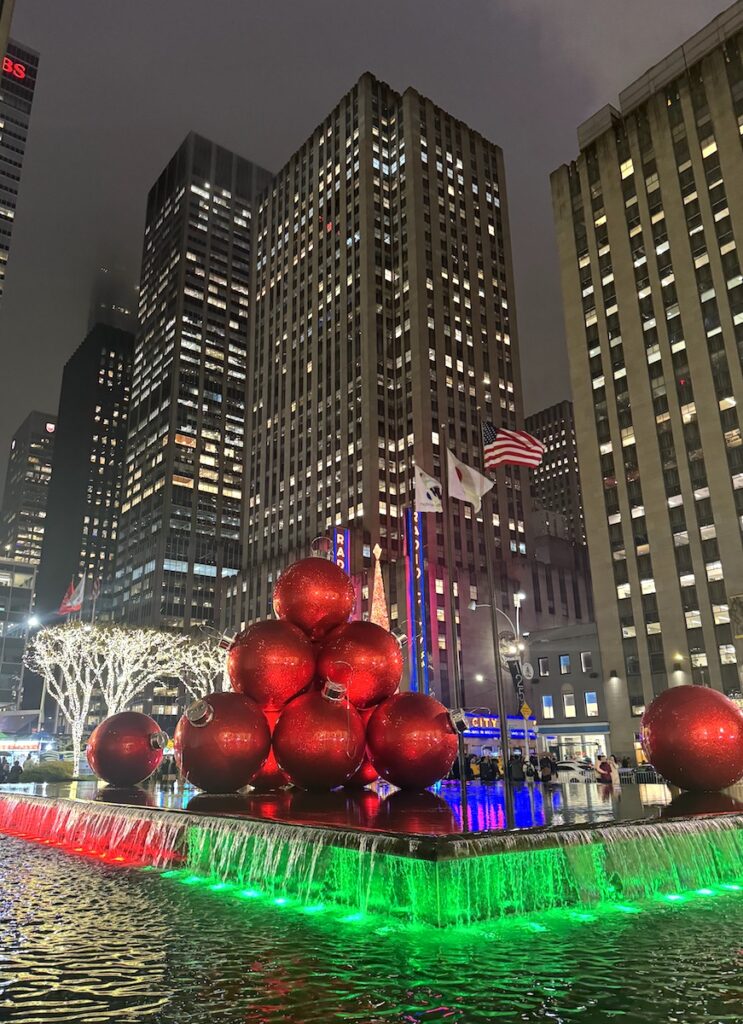 Giant ornament Christmas decorations in decorative fountain with large buildings rising up in background on winter night. Radio City Music Hall, Midtown Manhattan, New York City, New York.