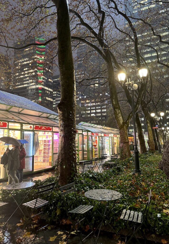 Christmas market in glass houses with large skyscraper buildings in background lit up at night, in foreground are people browsing the markets under umbrellas and rain covered bistro tables. Bryant Park Winter Village, Midtown Manhattan, New York City, New York.