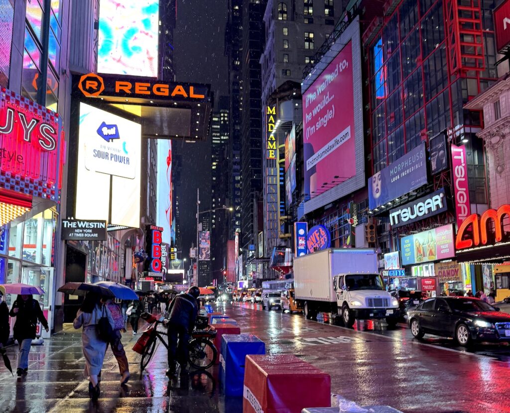 Downtown scene at night with brightly lit neon signs, huge billboards, and pedestrians and vehicle traffic moving through it on rainy night. Times Square, Midtown Manhattan, New York City, New York.