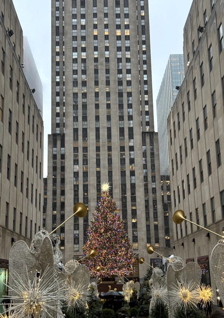 Large colorful Christmas tree in courtyard surrounded by huge towering buildings, walkway up to the tree is lined by giant decorations of angles playing trumpets and lit up stars. Rockefeller Center Christmas Tree, Midtown Manhattan, New York City, New York.