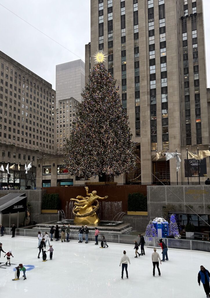 Ice skating rink under large colorful Christmas tree and golden statue surrounded by tall high rise buildings on gloomy afternoon. Ice Rink at Rockefeller Center, Rockefeller Christmas Tree, Midtown Manhattan, New York City, New York.