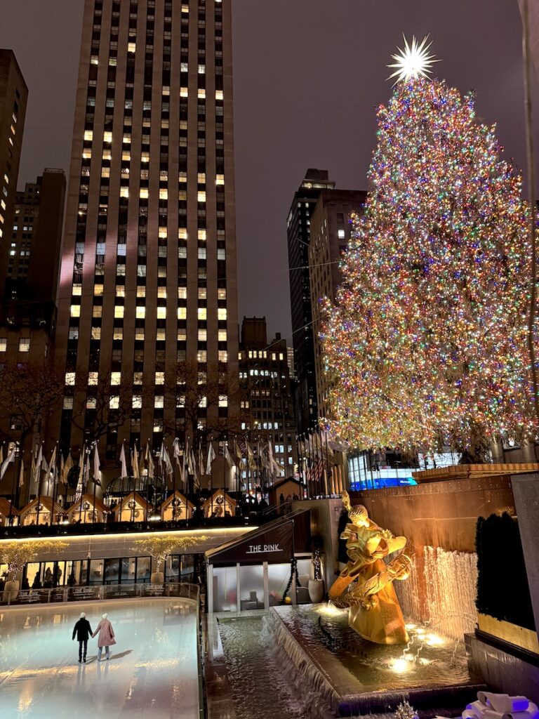 Couple holding hands and ice skating on rink under huge Christmas tree lit up at night. The Rink at Rockefeller Center & Rockefeller Christmas Tree. Midtown Manhattan, New York City, New York.