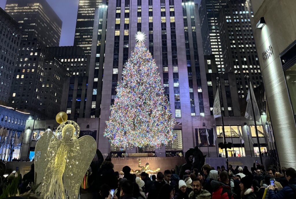 Crowds in front of huge Rockefeller Christmas tree lit up at night with towering skyscrapers in background. Rockefeller Center Christmas Tree, Midtown Manhattan, New York City, New York.