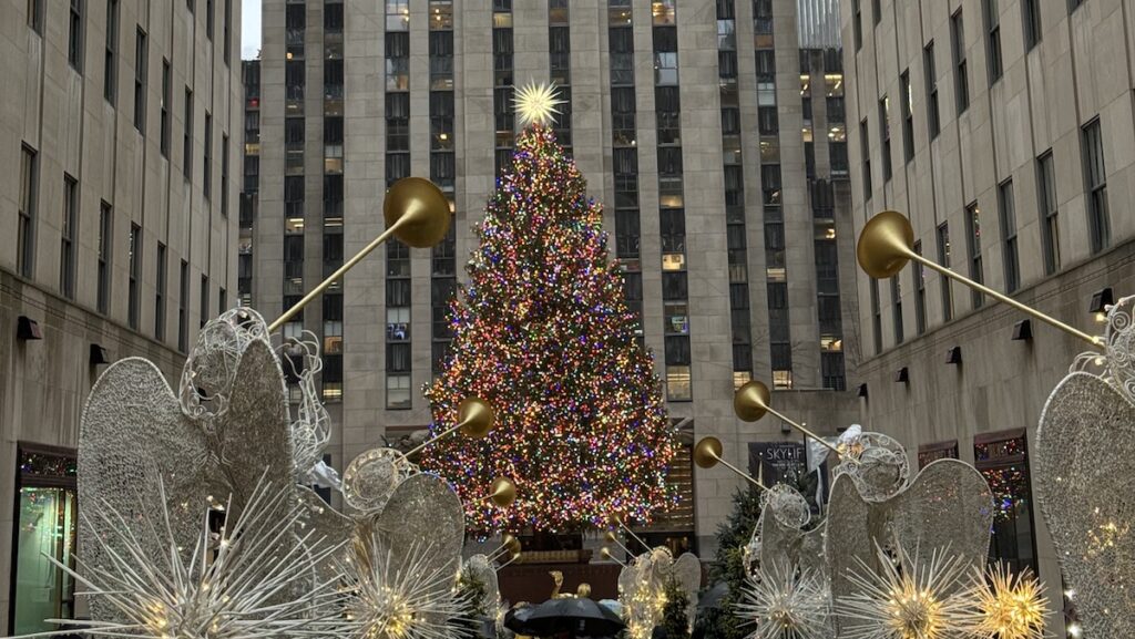 Large colorful Christmas tree surrounded by tall buildings with large angel statues holding trumpets in foreground. Rockefeller Center Christmas Tree, Midtown Manhattan, New York City, New York.