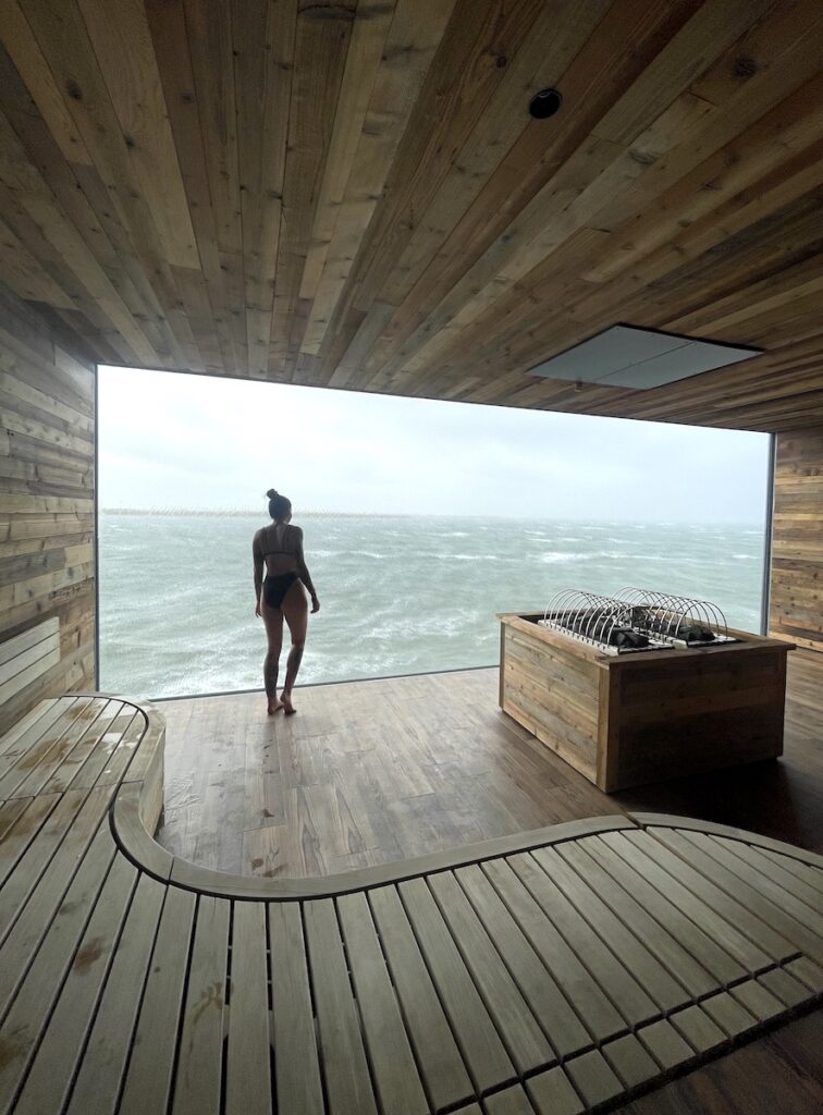 Woman in luxurious wooden sauna with large window overlooking ocean on rainy day. Sauna at Sky Lagoon, Reykjavik, Kópavogur, Iceland.