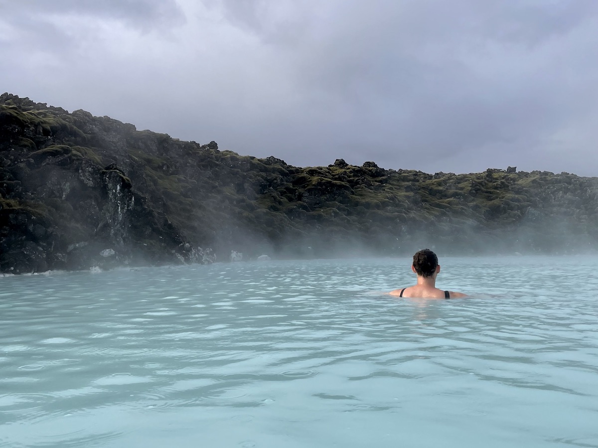 Woman soaking in mineral hot springs with bright blue water next to moss-covered lava field. Blue Lagoon, Grindavík, Iceland.