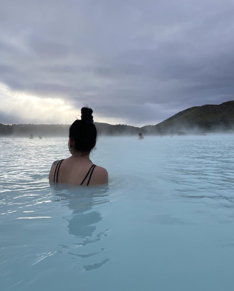 Woman soaking in hot spring while overlooking large lagoon of milky blue water on cloudy morning. Blue Lagoon, Grindavík, Iceland.
