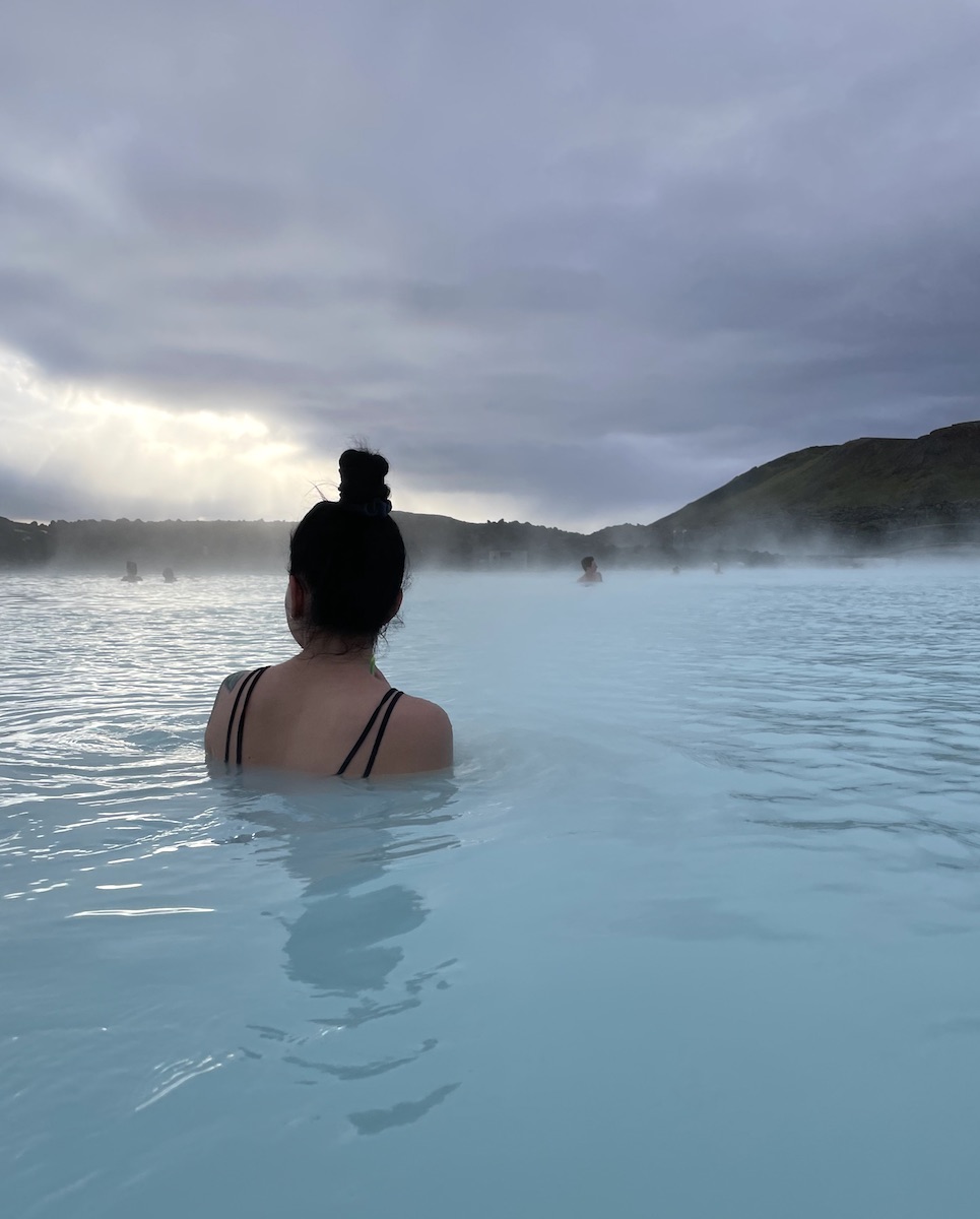 Woman soaking in hot spring while overlooking large lagoon of milky blue water on cloudy morning. Blue Lagoon, Grindavík, Iceland.