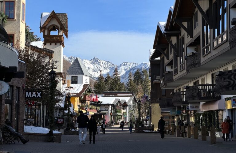 Vail Village lined with shops, restaurants, and Bavarian-style buildings with snow-peaked mountains in background on sunny winter day. Vail Village, Vail, Colorado.