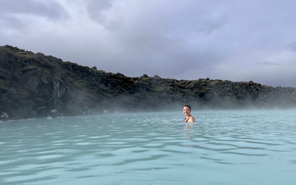 Woman smiling at camera while soaking in hot spring of bright blue water with moss-covered lava in background on sunny morning. Blue Lagoon, Grindavík, Iceland.